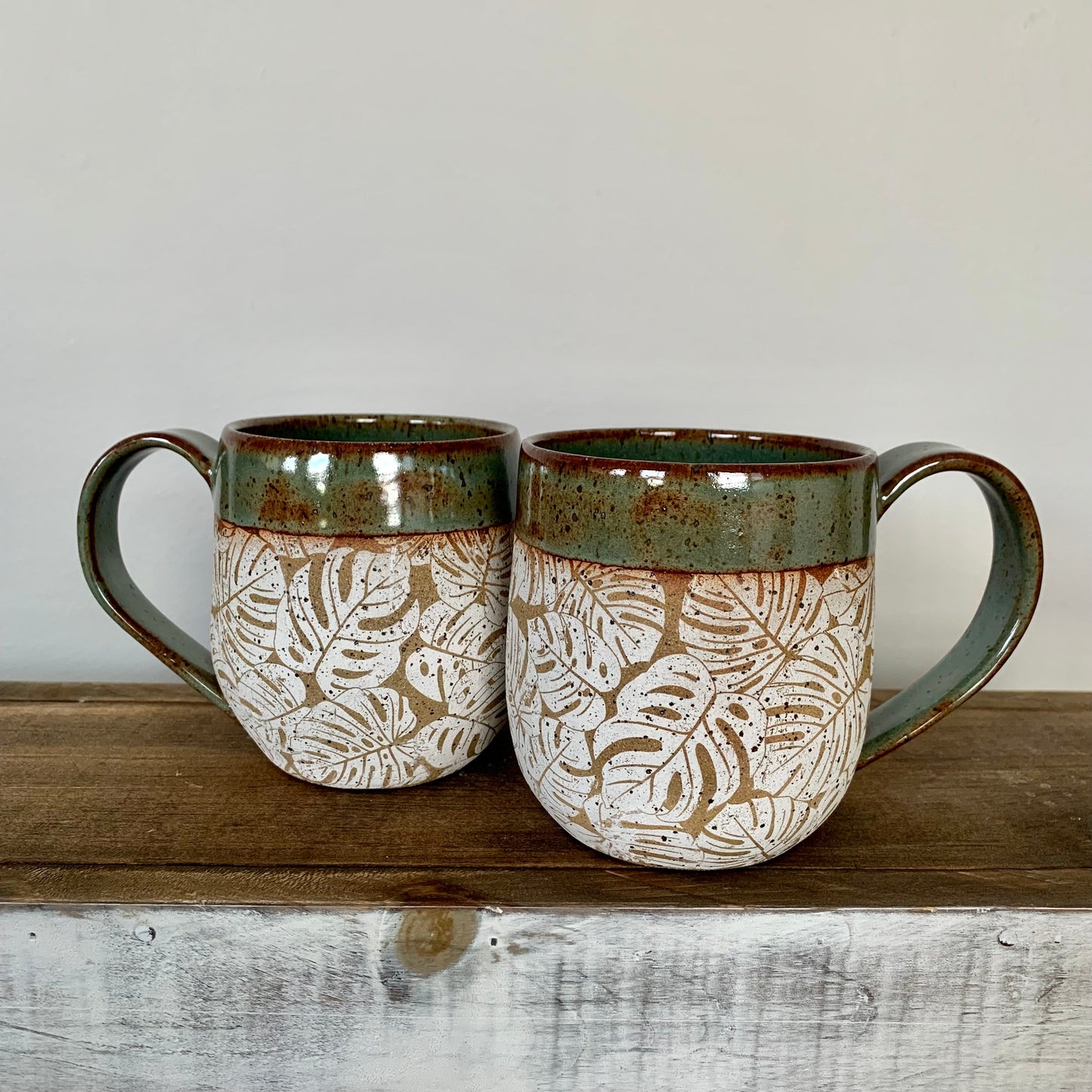 pottery monstera mug. The lush white  monstera leaves design wraps around the body of the mug which is natural brown speckled clay and accented with a nature green glaze on the handle and rim