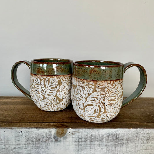 pottery monstera mug. The lush white  monstera leaves design wraps around the body of the mug which is natural brown speckled clay and accented with a nature green glaze on the handle and rim